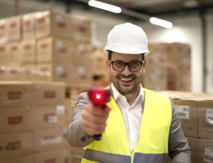 Portrait factory warehouse worker standing among cardboard boxes holding bar code scanner laser beam pointing camera - Operátor, Minőségellenőr, Mélyhúzó, Fröccsöntő